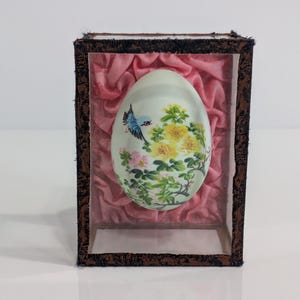 Puede incluir: Un objeto decorativo en forma de huevo con un diseño pintado de pájaro y flores, exhibido en una caja con frente de vidrio y respaldo de terciopelo rosa. La caja tiene un marco marrón oscuro.