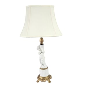 Pode incluir: Um abajur de mesa de porcelana branca com base dourada e cúpula de tecido branco. O abajur apresenta uma escultura de um querubim.