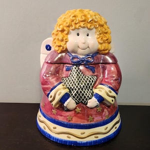 Può includere: Un barattolo per biscotti in ceramica a forma di angelo con capelli biondi ricci, che indossa un vestito rosso e blu e tiene una stella verde. Il barattolo ha uno sfondo bianco con stelle dorate e un bordo blu.