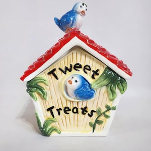 Pode incluir: Uma casa de pássaros de cerâmica em forma de casa de pássaros com um telhado vermelho e um pássaro azul no topo. A casa de pássaros tem uma placa que diz "Tweet Treats" e um pássaro azul no interior.