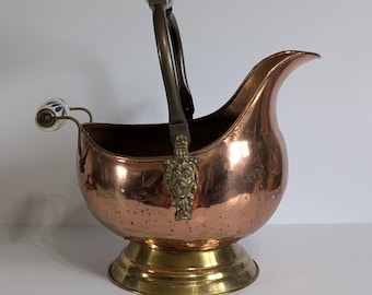 Può includere: Un secchio per carbone in rame con base e manico in ottone. Il manico presenta un emblema decorativo in ottone e dettagli in ceramica con motivi floreali blu. Il secchio ha un beccuccio curvo e una finitura lucida.