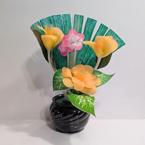 以下が含まれることがあります： 黄色とピンクの花、緑の葉、緑の扇形の背景が特徴の装飾的なフラワーアレンジメント。 花は光沢のある黒いセラミックの花瓶にアレンジされています。