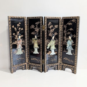 May include: A four-panel black lacquer screen featuring detailed illustrations of figures in traditional attire. The screen has gold accents and hinges, with each panel depicting a different scene. The figures are dressed in pastel colors.