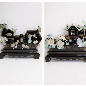 May include: Two black decorative wooden display pieces. Each features a black base with a detailed top, including a house, flowers, and birds. The designs incorporate white, blue, and gold accents, creating an ornate aesthetic. The pieces are set against a white background.