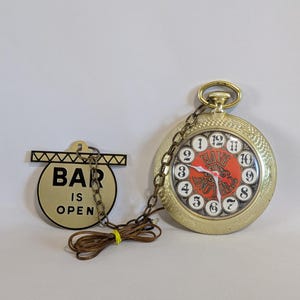 May include: A gold-colored pocket watch with a red face and black lettering that reads "Have Another". The watch is hanging from a chain and has a sign that reads "Bar is Open" hanging from the chain.