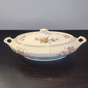 Puede incluir: Una fuente ovalada blanca con tapa, con un diseño floral en tonos naranja, azul y verde. La fuente tiene un borde dorado.
