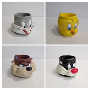 May include: Four ceramic mugs featuring Looney Tunes characters. The mugs include Bugs Bunny, Tweety Bird, Taz, and Sylvester. Each mug is designed in the shape of the character's head, with vibrant colours and detailed features.