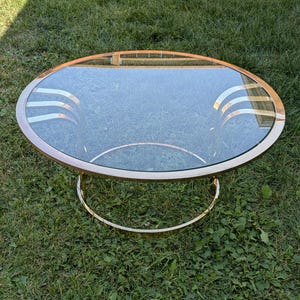 May include: A round coffee table featuring a clear glass top set within a polished gold-tone metal frame. The base consists of a circular metal ring connected to the tabletop by two sets of three curved, parallel gold-tone metal support bars.