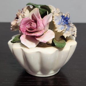 May include: A white ceramic flower pot with a fluted rim, filled with a bouquet of pink, white, and blue porcelain flowers. The flowers are detailed with green leaves and have a vintage aesthetic.