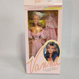 May include: A boxed "Limited Edition" Vanna doll featuring a blonde doll in a pink lace dress. The box has the name "Vanna" in pink script and an image of Vanna White. The box is pink and white.