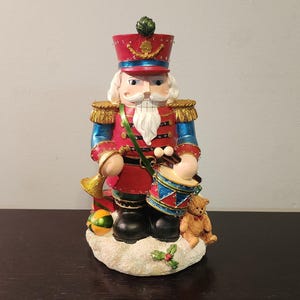 May include: A red and blue Christmas nutcracker figurine playing a drum with a teddy bear and a green and gold ball at its feet. The nutcracker is wearing a red and gold uniform with a blue hat and a white beard.