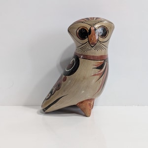 Op de afbeelding: Een keramieken uilfiguur met een beige lichaam, gedetailleerd met zwarte, bruine en rode accenten. De uil heeft grote, ronde ogen en een gestileerd ontwerp. Het beeldje staat op een wit oppervlak.