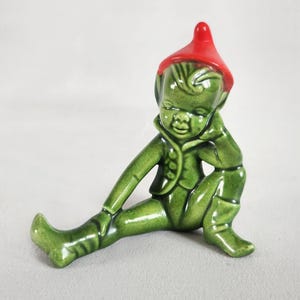 Peut inclure: Une figurine d'elfe en céramique verte avec un chapeau rouge, assise avec une jambe tendue et l'autre pliée, reposant son menton sur sa main.