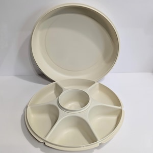Puede incluir: Un juego de bandejas de servicio de plástico blanquecino de dos piezas. La bandeja inferior está dividida en seis secciones, con un cuenco central. La bandeja superior es una tapa redonda simple. Diseñado para servir aperitivos.