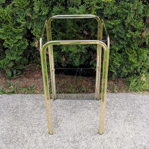 Op de afbeelding: Een hoge, rechthoekige standaard met een gepolijst goudkleurig metalen frame met afgeronde hoeken. De structuur bevat een heldere glazen plank nabij de basis, ondersteund door vier verticale metalen poten die een open frame vormen.