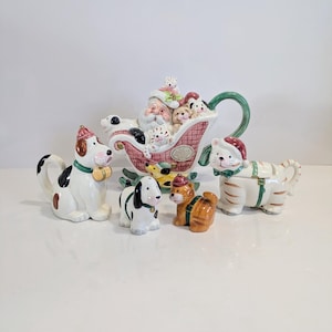 May include: A festive ceramic Christmas figurine set featuring Santa Claus in a sleigh pulled by dogs and cats. The figurines are brightly coloured with festive details, including hats and scarves. The set includes a teapot and individual figurines.