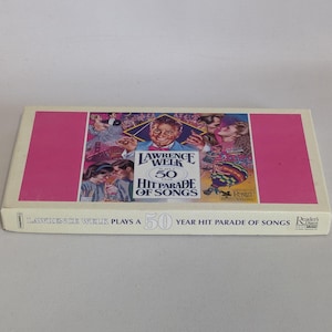 May include: A vintage cassette tape box for Lawrence Welk's "50 Year Hit Parade of Songs." The box has a pink border and a colourful image of Welk and other performers. The text "Lawrence Welk Plays a 50 Year Hit Parade of Songs" is printed on the box.
