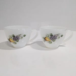Puede incluir: Dos tazas de té blancas con asas. Cada taza presenta un diseño de frutas con uvas, una pera y hojas verdes. Las tazas están hechas de un material translúcido y están sobre un fondo blanco.