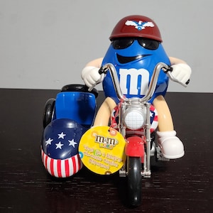 May include: A blue M&M's character in a helmet and sunglasses, on a red and silver motorcycle with a sidecar. The sidecar and helmet have patriotic designs. A yellow tag reads "M&M's World".