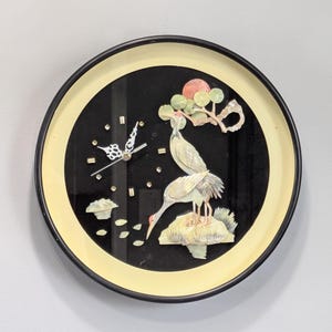 Puede incluir: Reloj de pared redondo con esfera negra y borde color crema. El reloj presenta una escena decorativa con dos grullas, un sol rojo y una rama de árbol. Las manecillas del reloj son plateadas y los marcadores de hora son pequeños cristales.