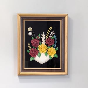 May include: A framed floral artwork featuring a white vase filled with red and yellow roses, set against a black background. The artwork is encased in a brown and gold frame, creating a vintage aesthetic.