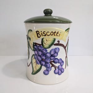 May include: A ceramic biscotti jar with a green lid and a decorative grape design. The jar has a cream-colored base with a banner that reads "Biscotti". The grapes are purple, and the leaves are green.