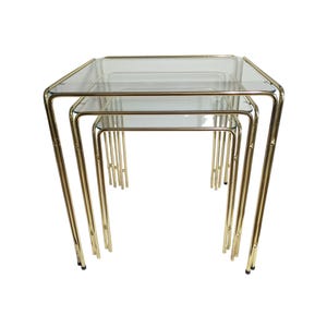 May include: Set of three nesting tables with clear glass tops and gold-colored metal frames. The tables are square and have a mid-century modern design. The legs are slender and slightly splayed, creating a stylish and functional furniture set.