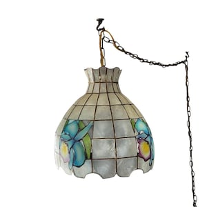 Op de afbeelding: Een witte, iriserende hanglamp met een bloemenmotief. De lampenkap is gemaakt van een raster van kleine, vierkante stukjes materiaal. Het bloemenmotief heeft blauwe, paarse en gele bloemen. De lampenkap hangt aan een ketting.