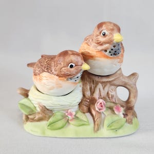 Pode incluir: Um conjunto de saleiro e pimenteiro de cerâmica com dois pássaros marrons empoleirados em um galho de árvore com um ninho. Os pássaros têm bicos amarelos e marcas marrons e brancas. A base é decorada com folhas verdes e flores rosas.