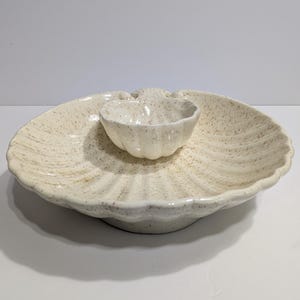 Puede incluir: Una fuente de cerámica color crema con un patrón moteado. La fuente tiene una bandeja exterior grande y festoneada y un cuenco más pequeño a juego en el centro. Diseñada para servir aperitivos o entremeses.