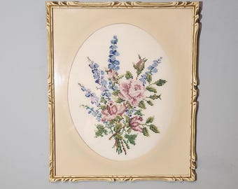 May include: A framed cross-stitch embroidery featuring a bouquet of pink roses and blue flowers in an oval shape. The frame is a light beige color with a decorative gold trim.