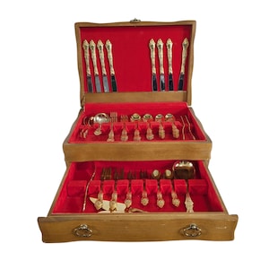 Może przedstawiać: Drewniana skrzynia na sztućce z czerwonym aksamitnym wnętrzem. Skrzynia jest otwarta, ukazując wiele warstw złotych sztućców, w tym noże, widelce i łyżki. Sztućce mają ozdobne uchwyty. Skrzynia ma drewnianą obudowę.