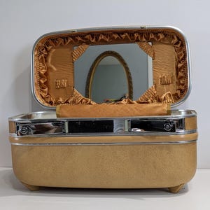 May include: Open vintage tan vanity case with a silver trim. The interior features a gold satin lining, a mirror, and a small handle. The case is rectangular with rounded corners and sits on small feet.
