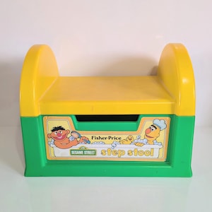 May include: A vintage Fisher-Price Sesame Street step stool. The step stool is primarily yellow and green, with a rectangular design. The front features a colourful graphic of Sesame Street characters and the words "step stool".