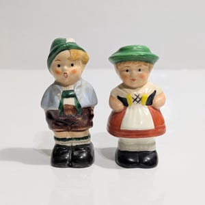 Könnte beinhalten: Zwei kleine Keramikfiguren von Kindern in traditioneller deutscher Tracht. Der Junge trägt einen grünen Hut, ein blaues Hemd und braune Shorts. Das Mädchen trägt einen grünen Hut, eine weiße Schürze und einen roten Rock. Beide haben schwarze Schuhe.