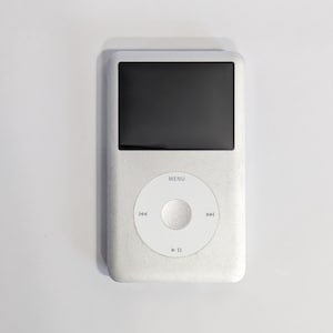 May include: Silver iPod with a black screen and a white control wheel. The control wheel has a menu button and forward, backward, and play/pause buttons. The iPod is on a white background.