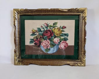 May include: A framed needlepoint artwork featuring a bouquet of roses in shades of red, pink, and yellow, arranged in a blue vase. The artwork is set against a beige background with a green border, encased in a gold-colored, ornate frame.