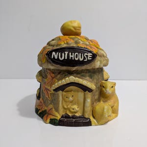 Peut inclure: Pot à biscuits en céramique "NUTHOUSE" en forme de chalet forestier. Le pot présente un écureuil jaune, des feuilles d'automne et une petite porte avec des figurines d'écureuils. Le couvercle est surmonté d'un gland jaune.