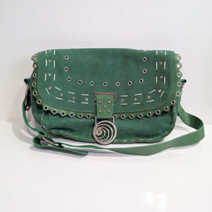 May include: Green suede shoulder bag with silver-tone hardware. The bag has a flap closure with a decorative buckle, eyelet and stitch detailing, and a long adjustable strap. The bag is a medium size.