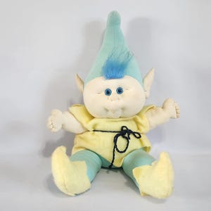 Puede incluir: Un duende de peluche con pelo azul y un atuendo amarillo y azul. El duende tiene grandes ojos azules y una amplia sonrisa.