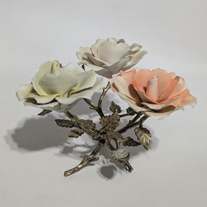 May include: A decorative floral arrangement featuring three ceramic roses in shades of cream, peach, and pale yellow. The roses are mounted on a bronze-colored metal base with leaves and stems. The arrangement is a decorative accent piece.