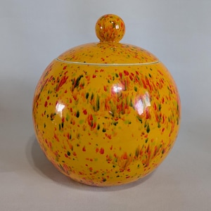 May include: A spherical, yellow ceramic jar with a lid and a small spherical handle. The jar is decorated with a speckled pattern of red, green, and black dots. The lid has a white rim.