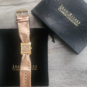 May include: A gold-toned watch with a rose gold leather strap and a square face. The watch is in a black velvet pouch with the words "Joan Rivers Classics Collection" on the pouch.
