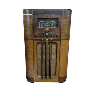 Può includere: Una vecchia radio in legno con la parte superiore marrone scuro e i lati marrone chiaro. La radio ha un display rettangolare con un quadrante verde e giallo. La parte anteriore presenta una griglia per l'altoparlante e diverse manopole.