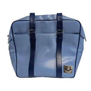 May include: A light blue Roxy handbag with navy blue straps and accents. The bag features a rectangular shape with a textured surface and a gold-colored Roxy logo on the front. The bag has two navy blue handles.