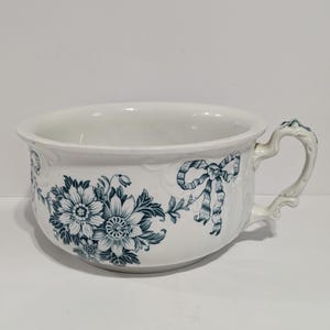 Puede incluir: Cuenco de cerámica blanca con asa, decorado con motivos florales y lazos color turquesa. El cuenco tiene un borde ancho y un asa curvada con detalles decorativos. El diseño floral incluye flores y hojas grandes y detalladas.