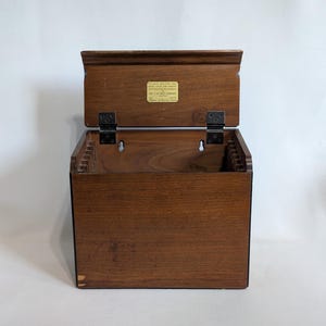 May include: A wooden recipe box with the lid open, revealing interior dividers. The box is a warm brown color with a rectangular shape and a hinged lid. A small label is visible on the inside of the lid.