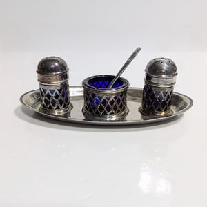 May include: A silver salt and pepper set with a matching open salt cellar and spoon, presented on an oval tray. The set features a lattice design over cobalt blue glass inserts. The salt and pepper shakers have silver lids.