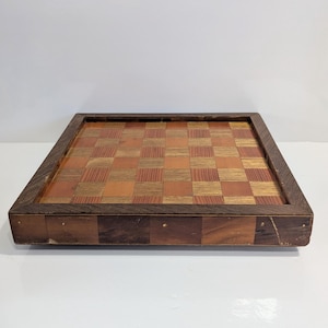 May include: A vintage wooden chessboard with a dark brown frame. The playing surface features a checkerboard pattern of alternating light and dark wood squares. The board is square and appears to be well-crafted.