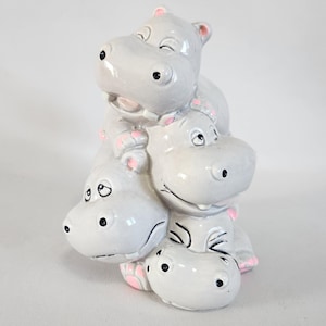 Pode incluir: Uma escultura de cerâmica de quatro hipopótamos cinzentos empilhados uns sobre os outros. Os hipopótamos têm detalhes cor-de-rosa nas orelhas e nos pés, e olhos pretos. Cada hipopótamo tem uma expressão sorridente.